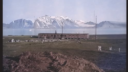 Photo of a single-story, elongated building, with water and a mountain partially covered with snow in the background. Information about the exhibition at the bottom of the photo.