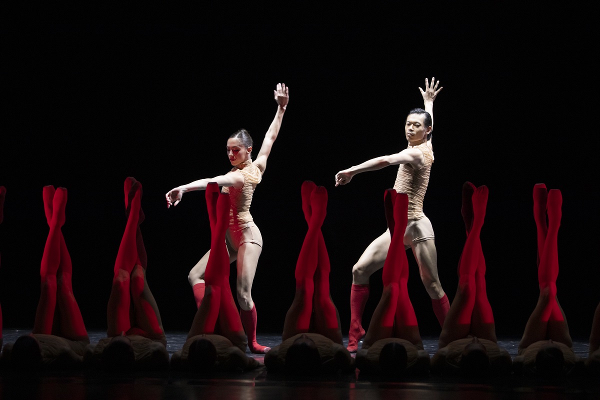 Two dancers illuminated by the light on the stage, in front of them dancers lying on the floor with their straight legs raised up in red tights. - grafika artykułu