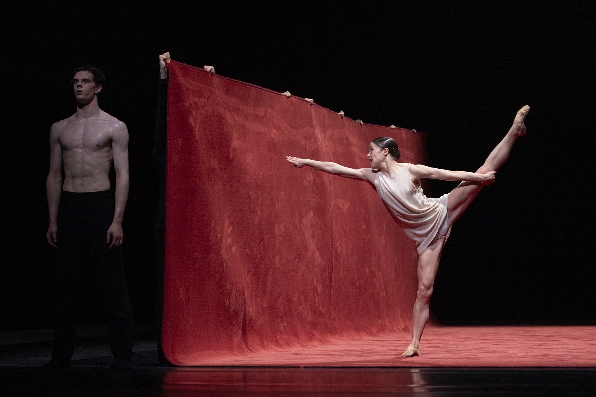 A ballerina in a dance pose in front of a red vertical plane. Behind the red plane a male ballet dancer standing straight. - grafika artykułu