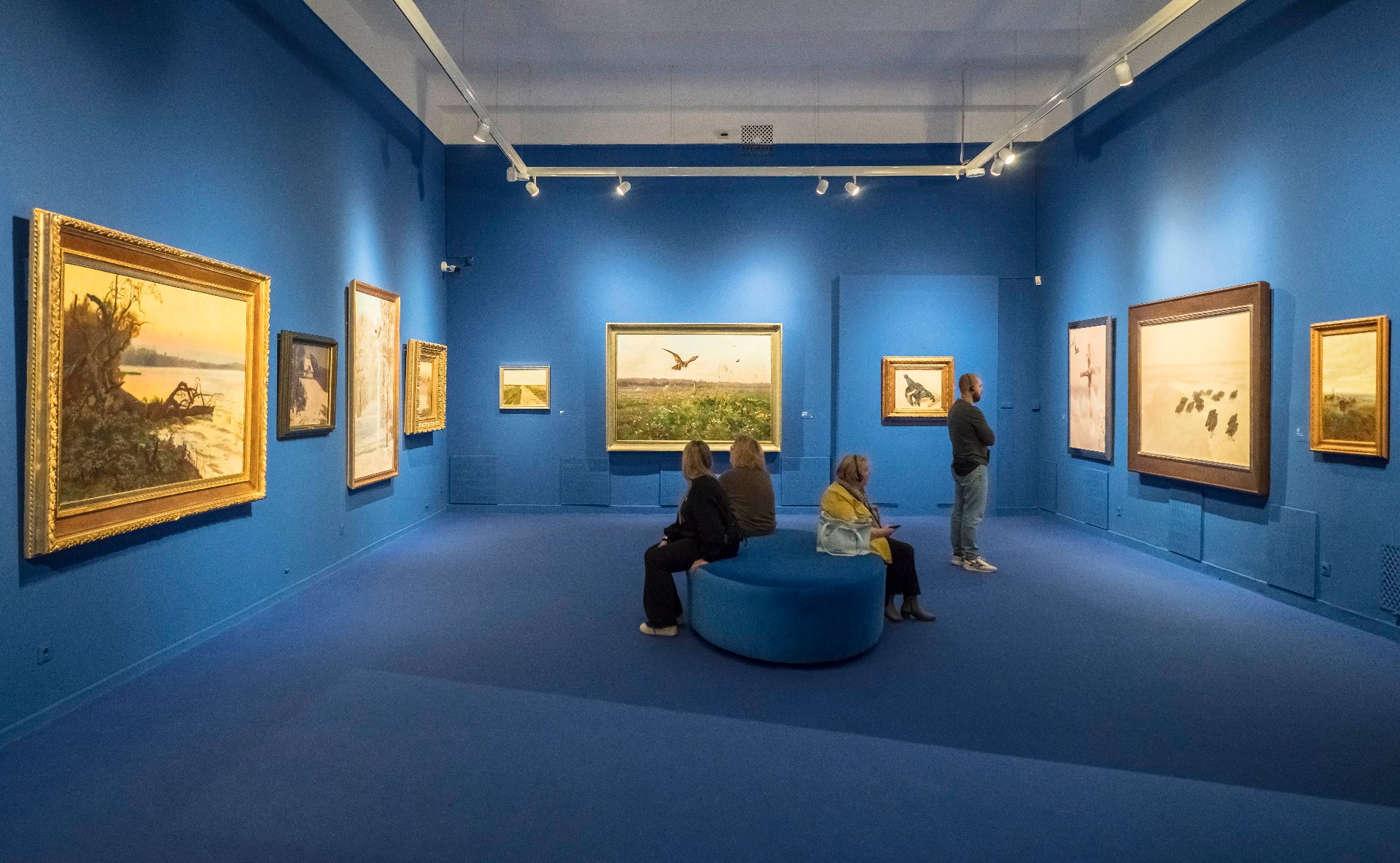 Exhibition hall. On three blue walls there are paintings of various sizes in frames. In the center there is a large round pouf on which three people are sitting. - grafika artykułu