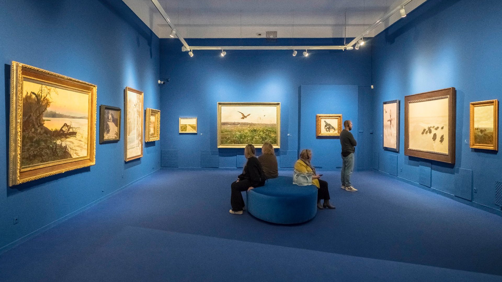 Exhibition hall. On three blue walls there are paintings of various sizes in frames. In the center there is a large round pouf on which three people are sitting.