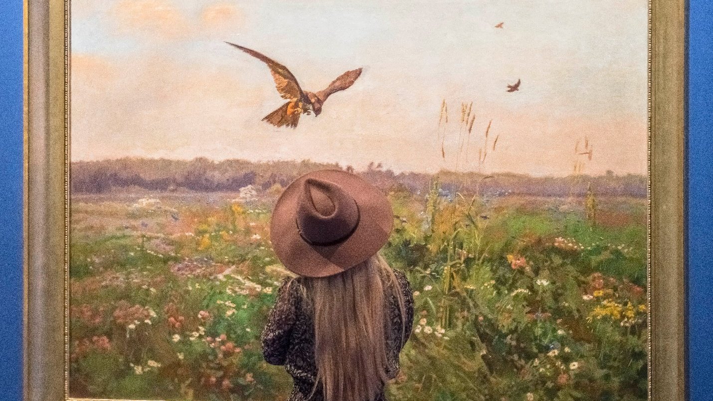 A woman is looking at a painting. We see her back, she is wearing a hat. In the painting, a bird of prey flies over a field.