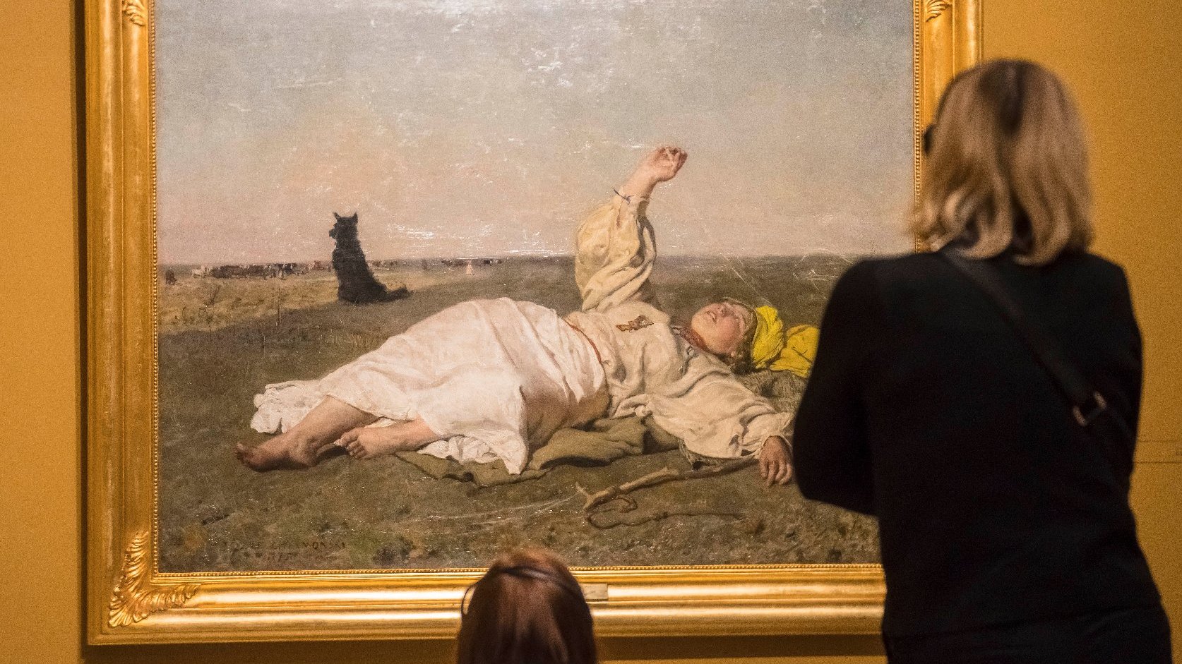 Two people are looking at a painting, we see their backs. In the painting in a gold frame, a figure is lying on the ground with one hand raised, in the background a black dog.