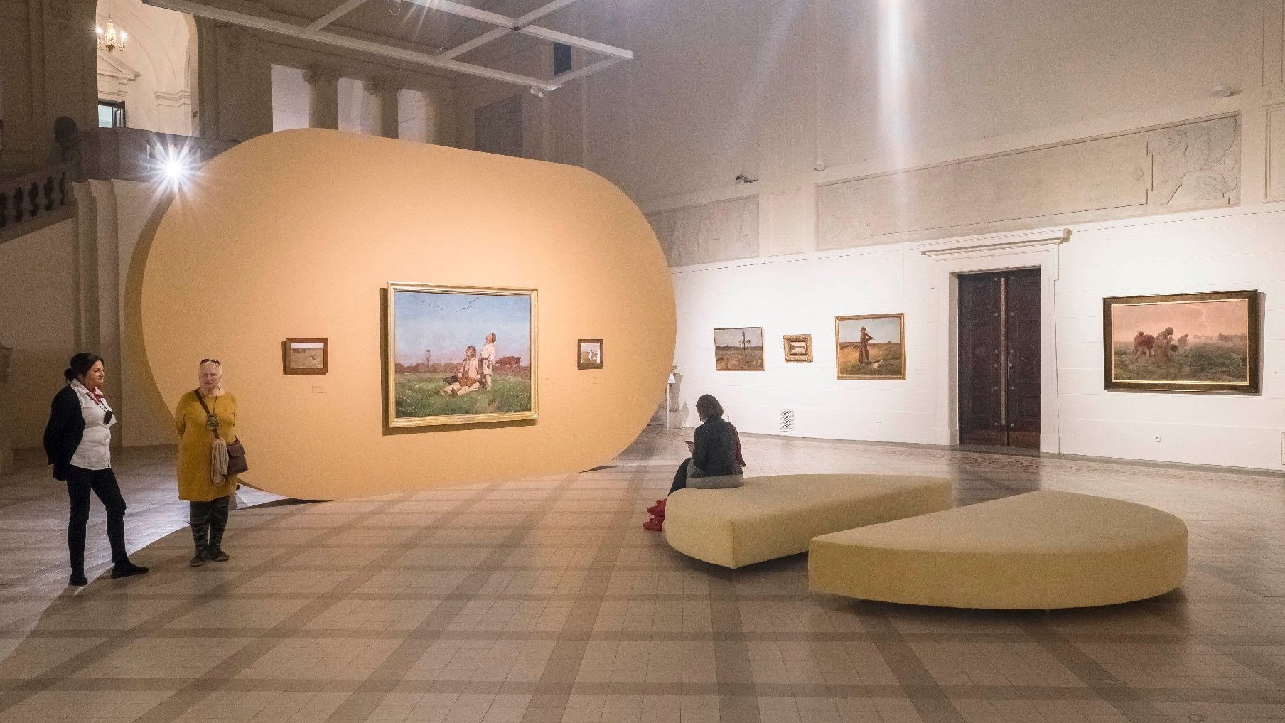 Exhibition hall, paintings on the walls, high ceiling. In the middle is a pouf - two semicircles. A woman is sitting on it. Two other women are looking at paintings.
