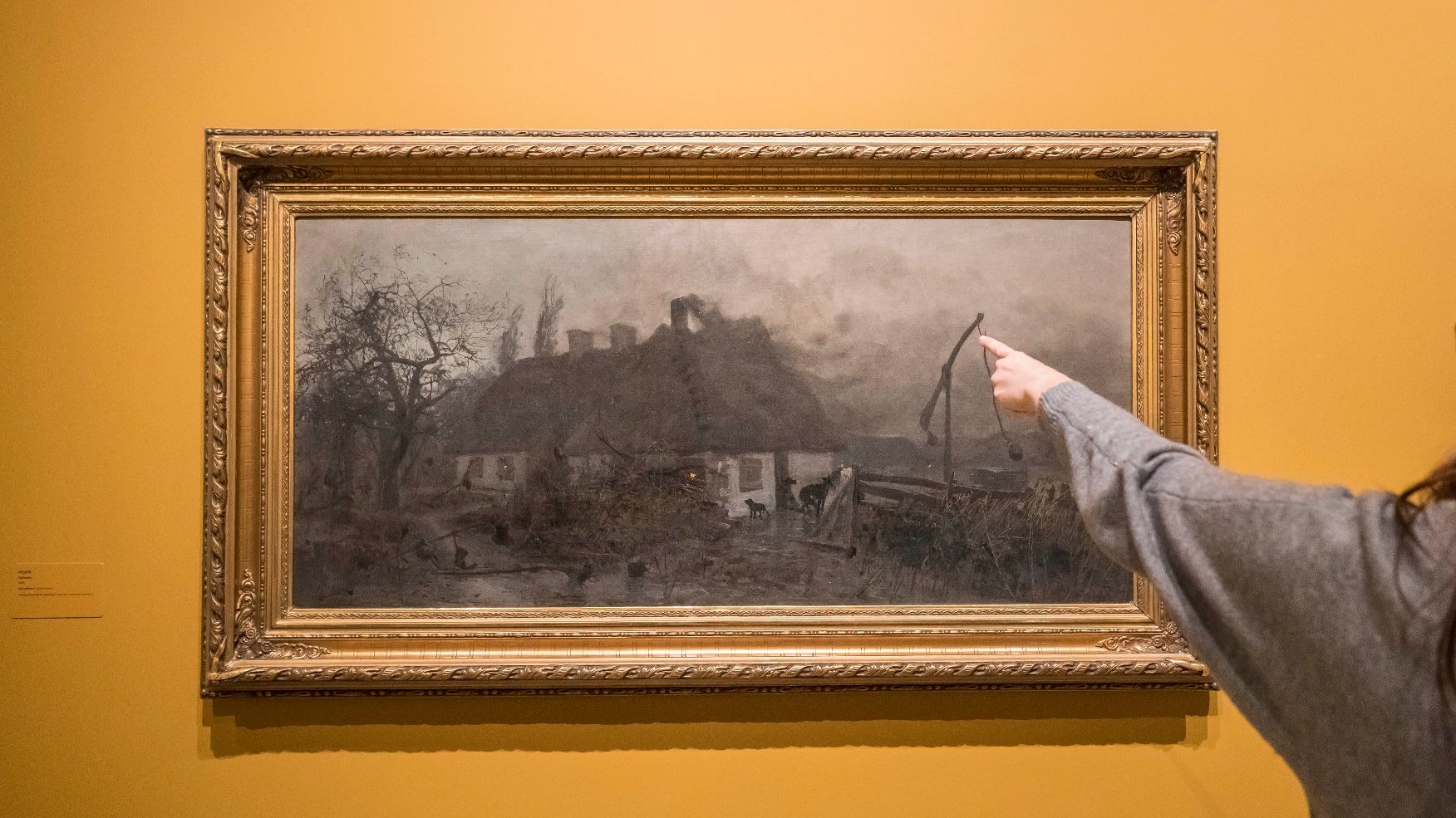 A picture in a gold frame hanging on the wall. An old cottage is depicted in the picture. Someone is pointing at an element of the picture with a finger.