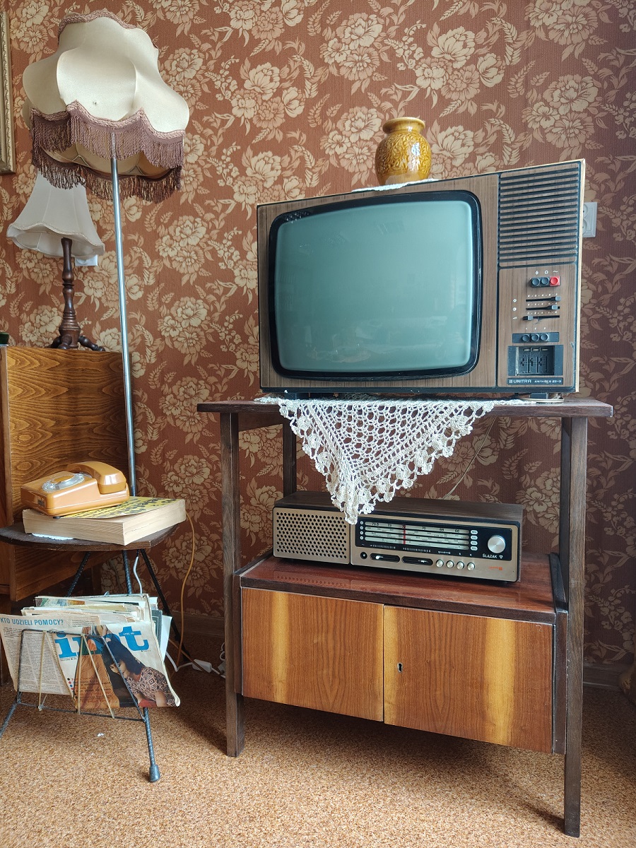 An old television set standing on a small table, under it old-fashioned radio. On the left a lamp and a small stand on which a dial telephone and a phone book are lying. Patterned white and brown wallpaper as a background. - grafika artykułu