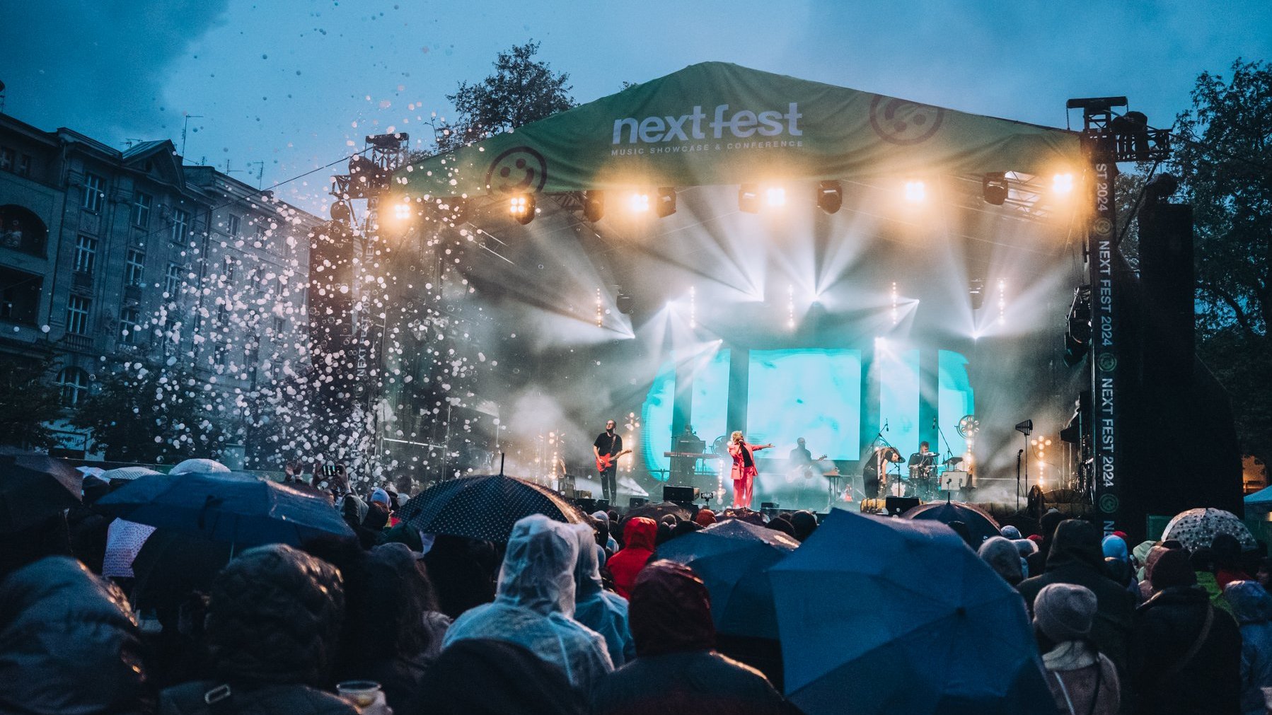 A lit stage with a band and a singer, and a crowd of people with umbrellas in front of it
