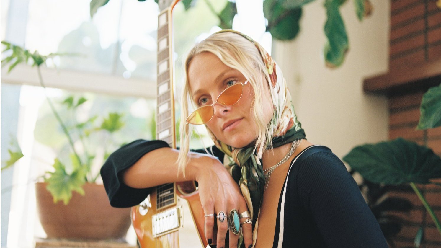 A sitting, thoughtful woman holds an electric guitar with the neck facing up.