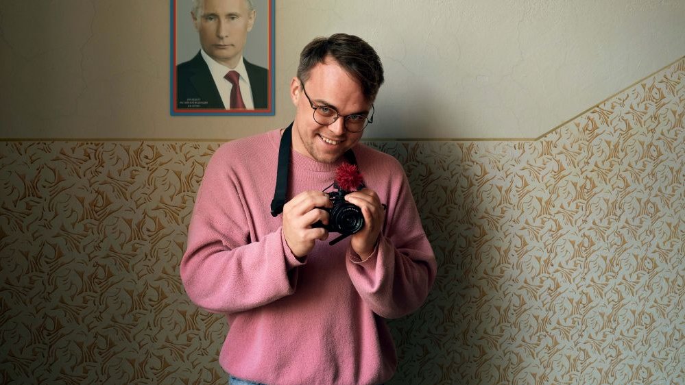 A smiling young man holds a camera in his hand. Behind him, on the wall, hangs a portrait of Putin.
