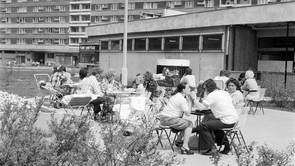 Black and white photo of several tables and people sitting at them on a concrete terrace. A block of flats in the background.