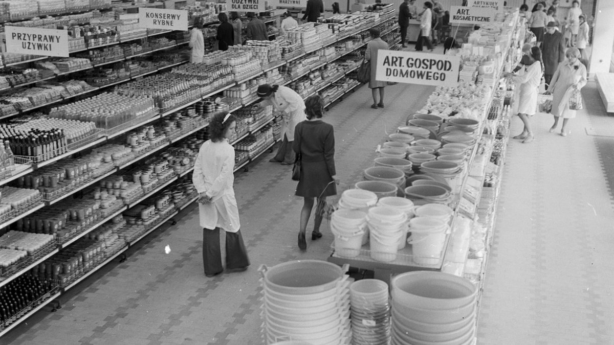 A black and white photo of the interior of a supermarket, with several staff members and shoppers.