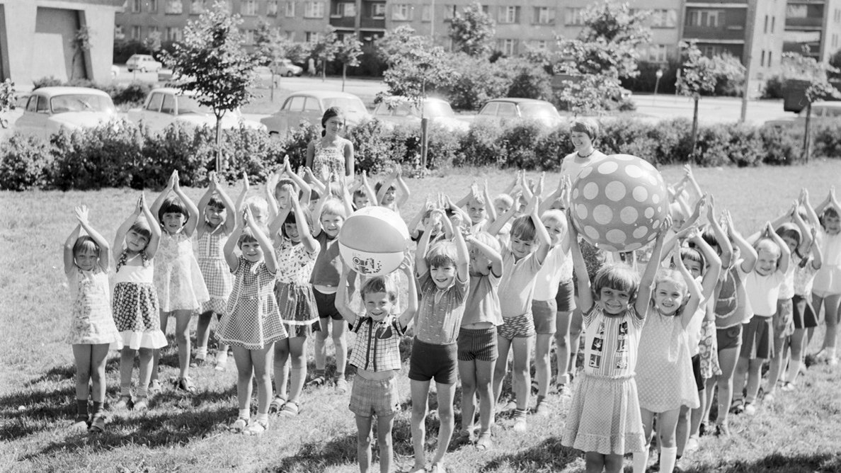 Black and white photo of children standing in four rows on grass, with their hands up. Two kids are holding big beach balls. A residential buildings in the background.