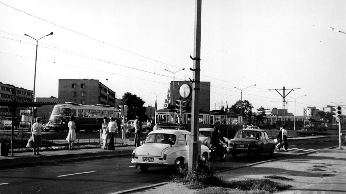 Black and white photo of a street with cars, trams and a bus from 1970s. Some people standing an a tram stop.