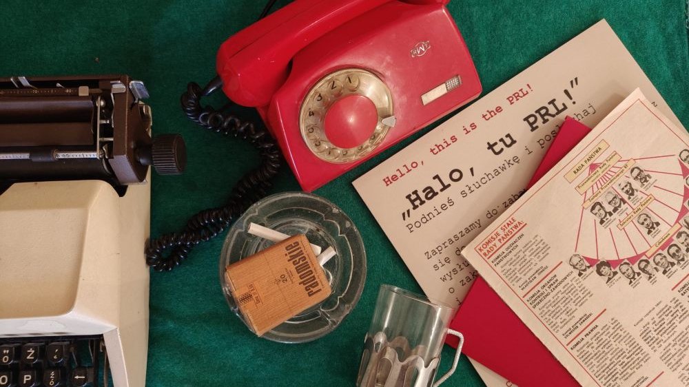 On a dark green tablecloth, several items from the PRL era, such as a telephone, a typewriter, a glass and an ashtray.