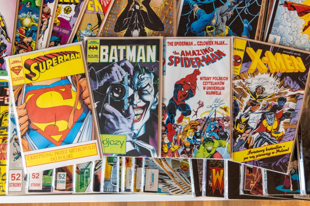 Four full color covers of Superman, Batman, Spider-Man, and X-men comics. They lie on a stack of many other comics. - grafika artykułu