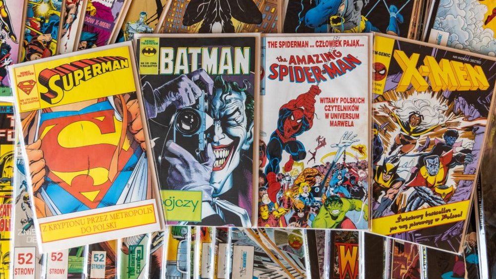 Four full color covers of Superman, Batman, Spider-Man, and X-men comics. They lie on a stack of many other comics.