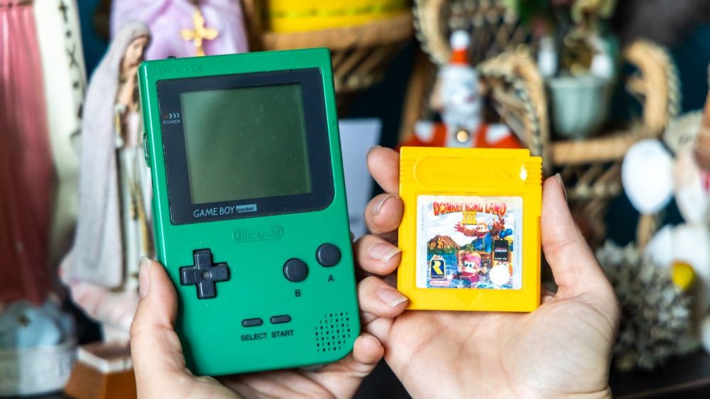 The hands hold a green, old Gameboy with a screen, next to it is a small yellow diskette with a game.
