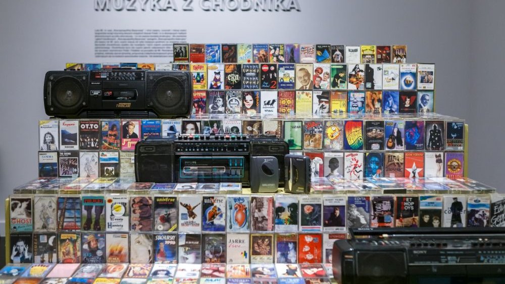 On several shelves, there are many cassette tapes, three black tape recorders and two walkmans. A slogan written on the wall: "Muzyka z chodnika"