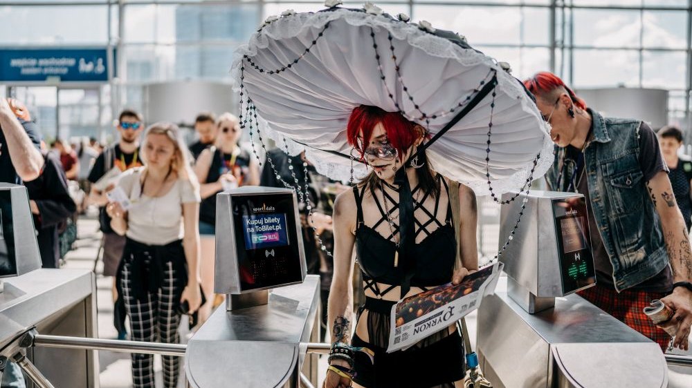 A red-haired girl in a large, white hat resembling a mushroom passes through the gates at Poznań International Fair