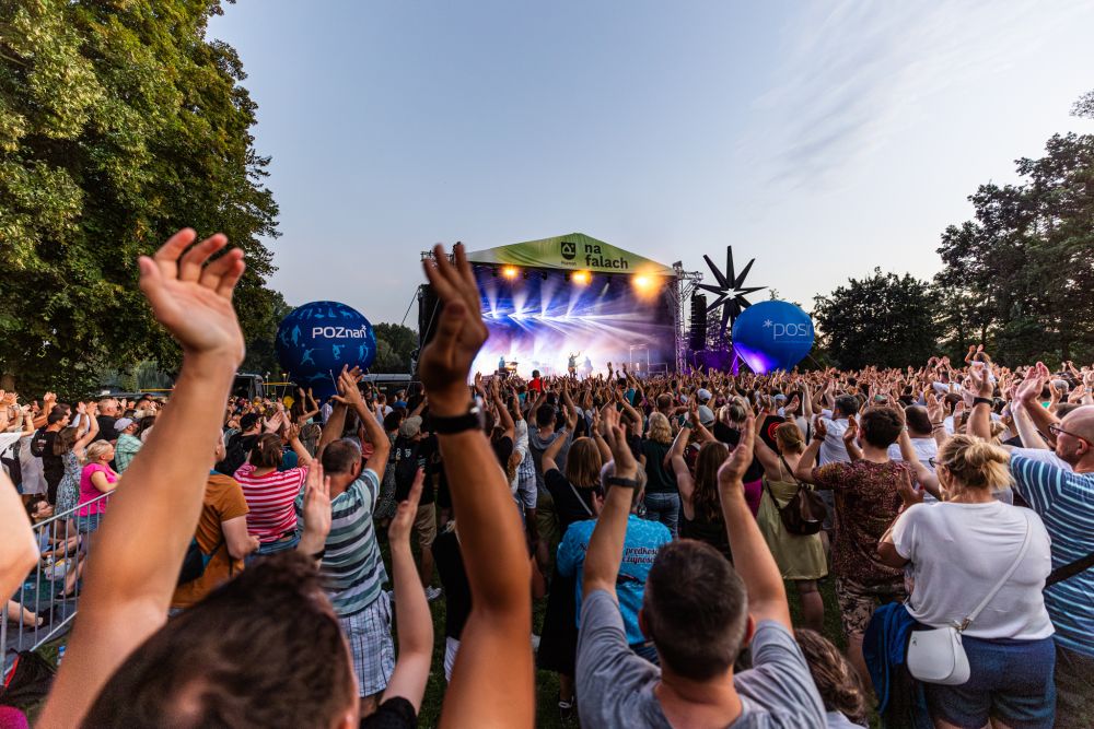 A bright outdoor stage with several hundred people partying in front of it. Many of them clap their hands over their heads. - grafika artykułu