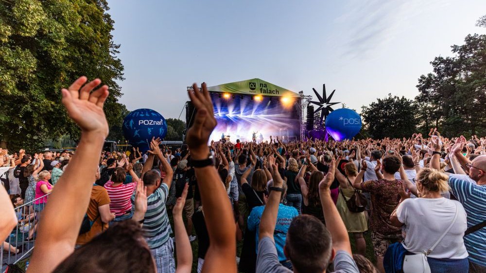 A bright outdoor stage with several hundred people partying in front of it. Many of them clap their hands over their heads.