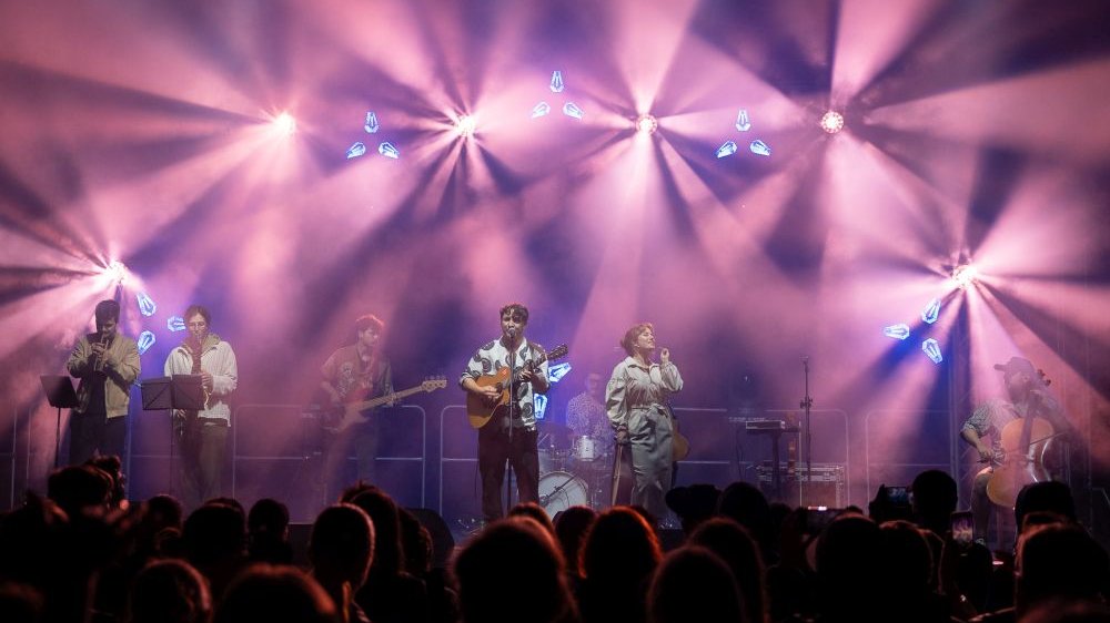 The stage is lit up in pink, a band is performing on it. In the center there is a vocalist who plays a classical guitar.