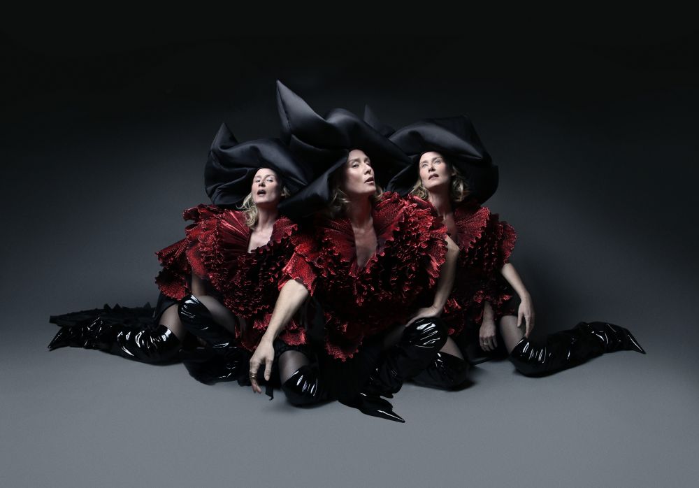 Three women are sitting on the floor. They are wearing burgundy dresses, black high boots, and black hats. Dark gray background. - grafika artykułu