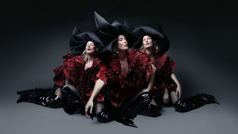 Three women are sitting on the floor. They are wearing burgundy dresses, black high boots, and black hats. Dark gray background.