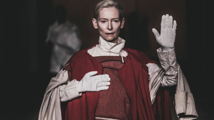 The actress in a burgundy and white robe and white gloves stands with one hand raised, the other held on her chest. Dark background.