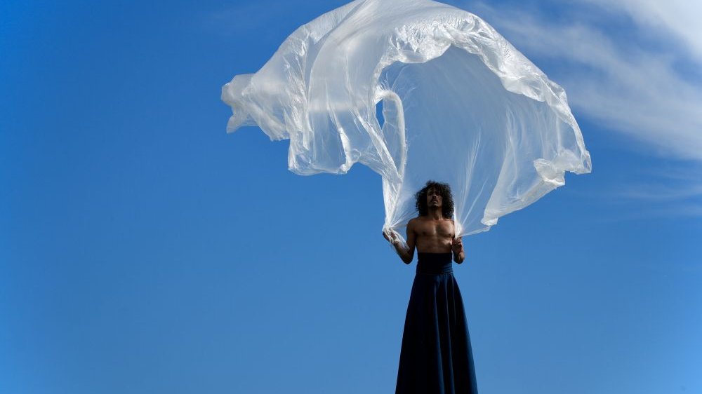 A man in a long navy blue skirt holds above himself a white foil resembling a parachute canopy. Blue sky as a background.
