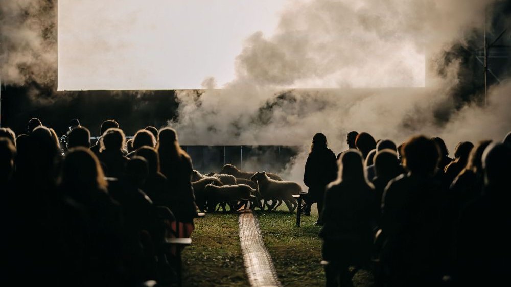 Several rows of spectators, sheep passing in front of them. Smoke rising in the background.