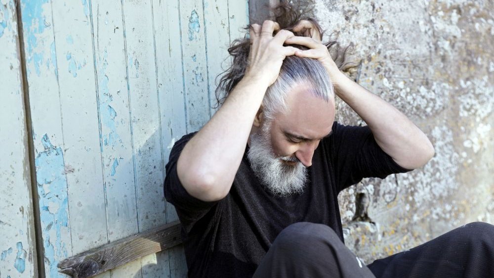 The man sits leaning against an old light blue wooden door, his head lowered and his hands holding his hair back.