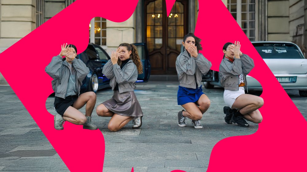 Four girls in gray jackets and short skirts crouch down and cover their faces with their hands. A building is in the background.