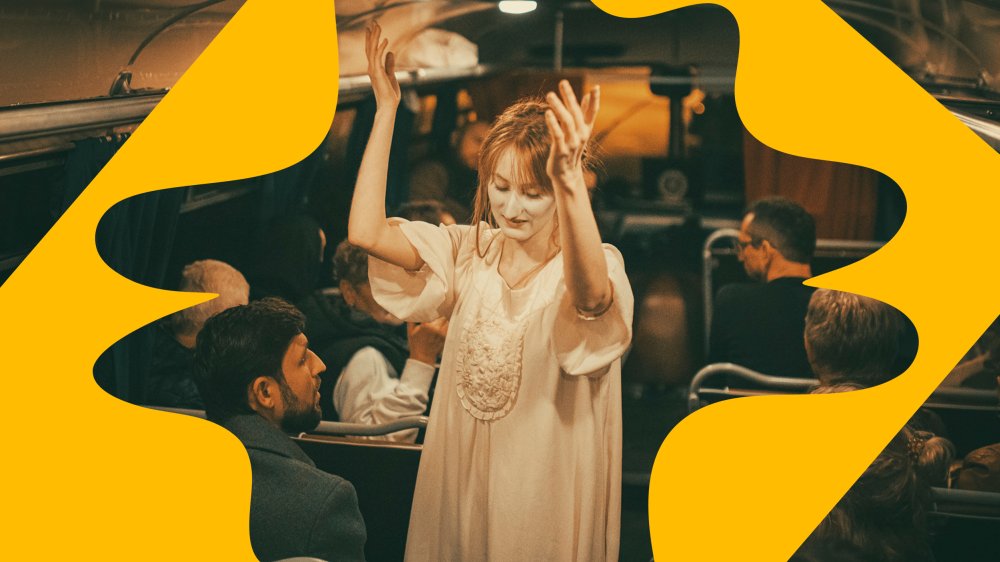 A woman in a long, light dress with her arms raised, behind her several people sitting on the bus with their backs to her.