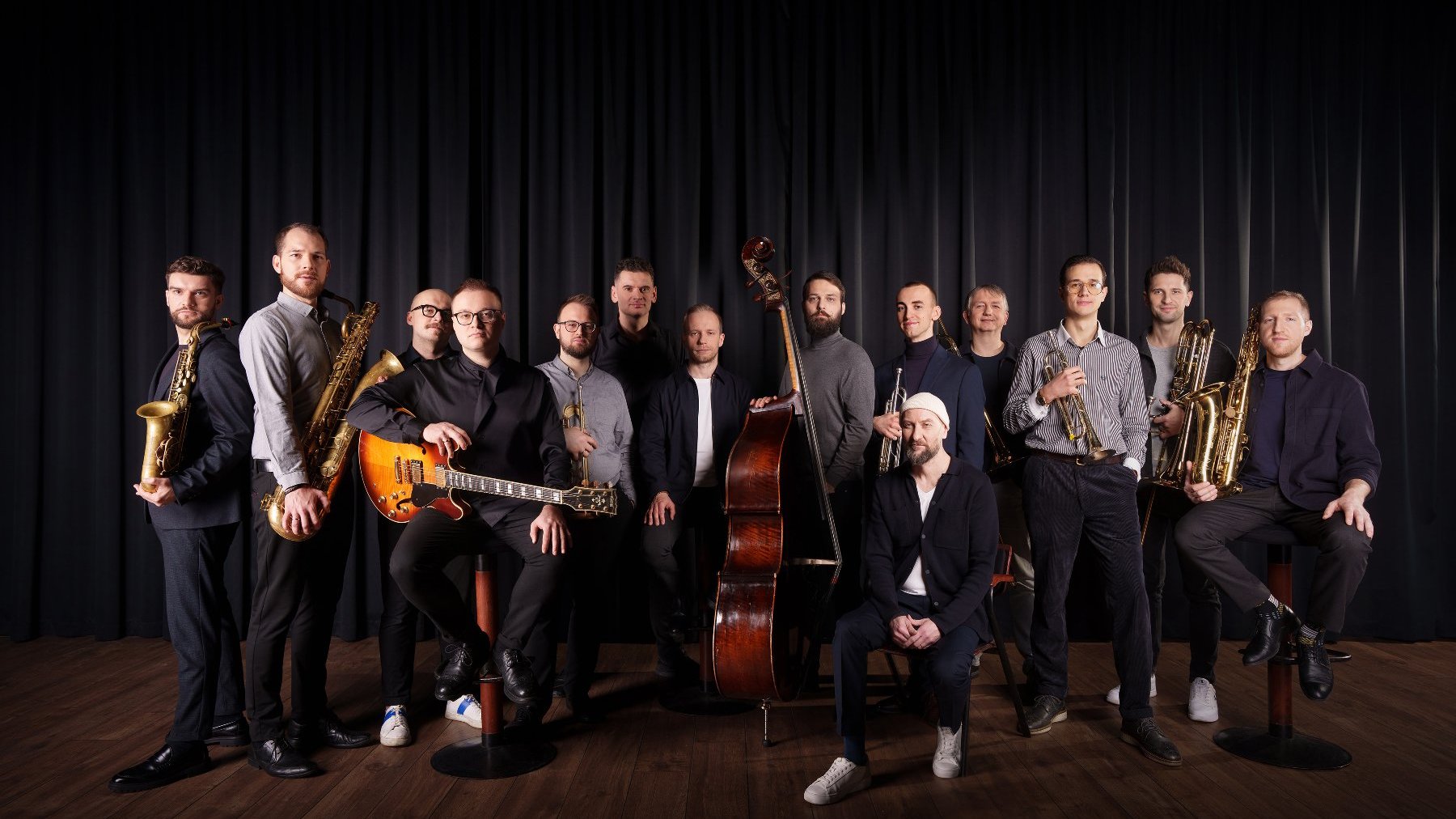 Several musicians, some of them standing, some sitting; a few of them are holding musical instruments such as a trumpet, a guitar or a saxophone.