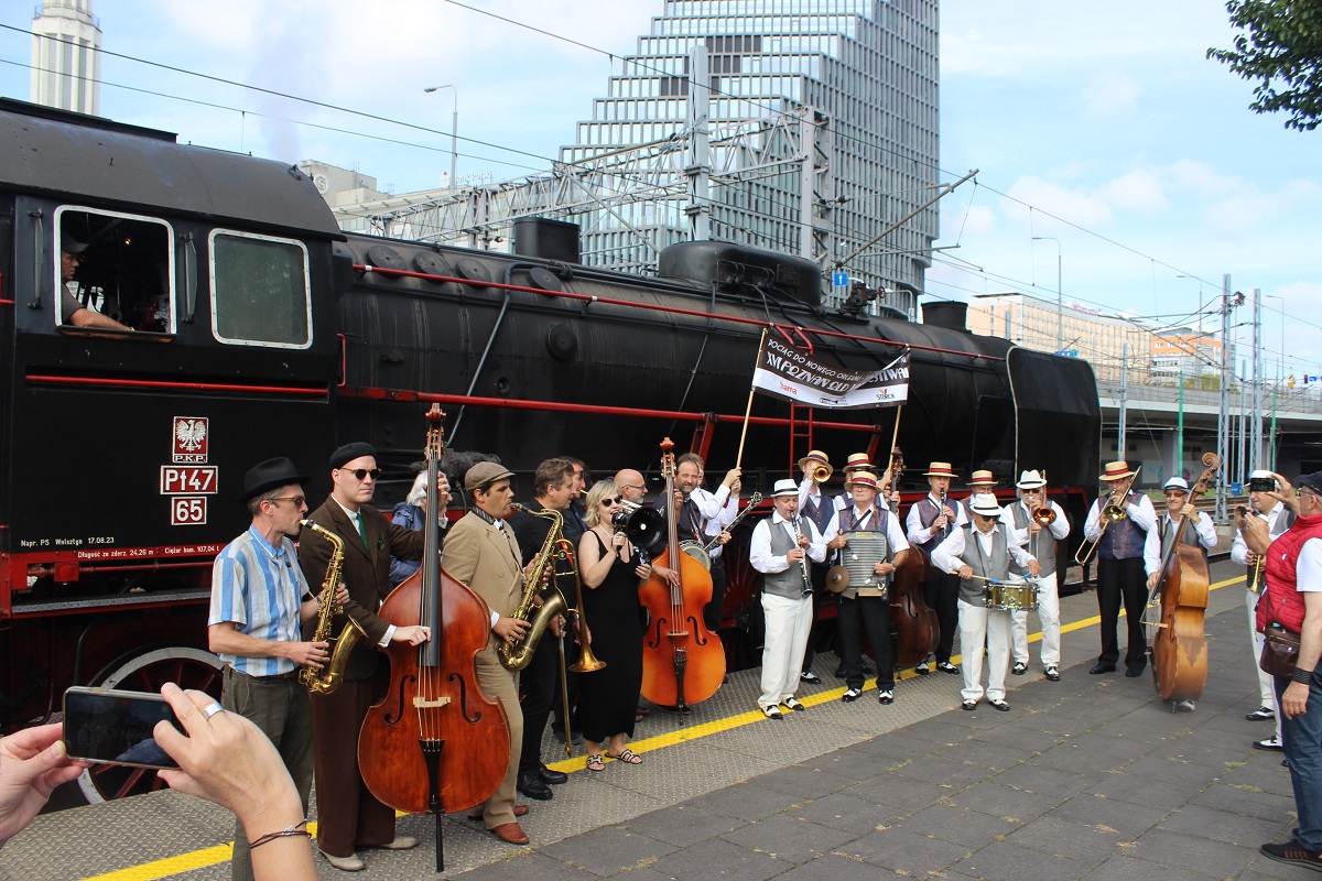 A group of musicians with musical instruments standing on a railway platform by the steam locomotive. - grafika artykułu