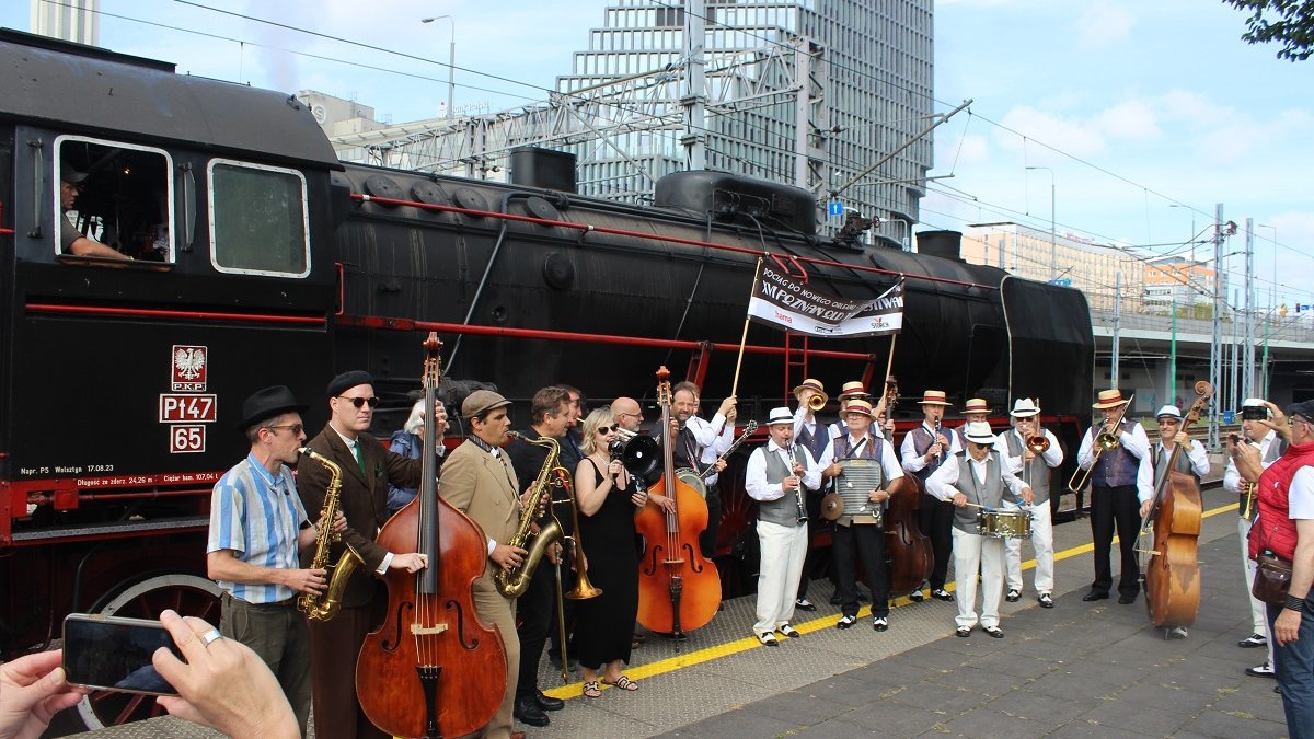 A group of musicians with musical instruments standing on a railway platform by the steam locomotive.