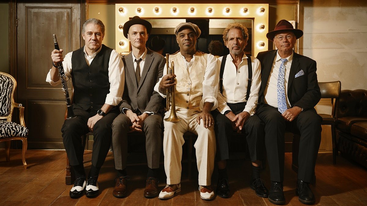 Five musicians sitting on a bench, two of them are holding musical instruments. In the background a mirror with bulbs.