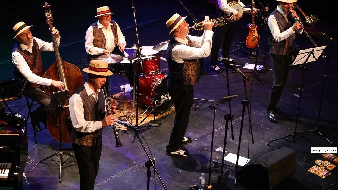 Jazz band performing on stage: six men wearing hats, white shirts and vests and playing various instruments.