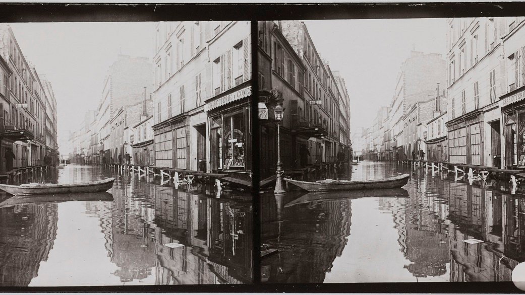 Stereophotograph showing a flooded street with buildings on both sides of it. An empty boat in the midle of a flooded street.