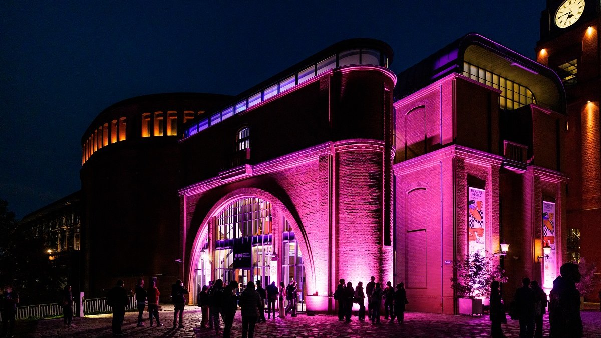 The Old Brewery building at night, illuminated by purple light. Small groups of people in front of the building.