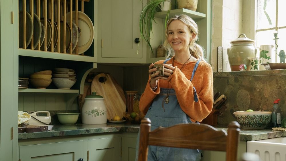 A smiling girl stands with a mug in her hands, leaning against the kitchen counter. There are kitchen cabinets and a small window behind her.