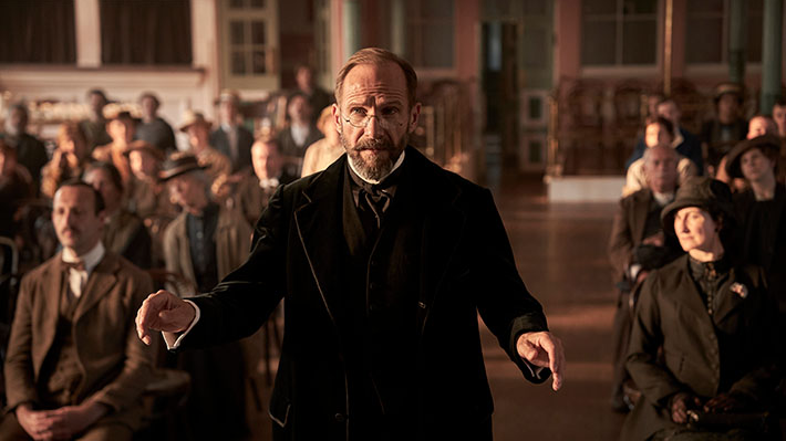 A man in old-fashioned clothes stands with his back to the seated people.