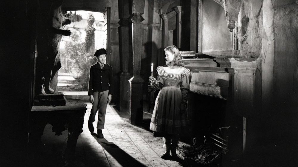 A black-and-white photograph of a girl and a boy standing in a dark room. The girl is holding a candle.