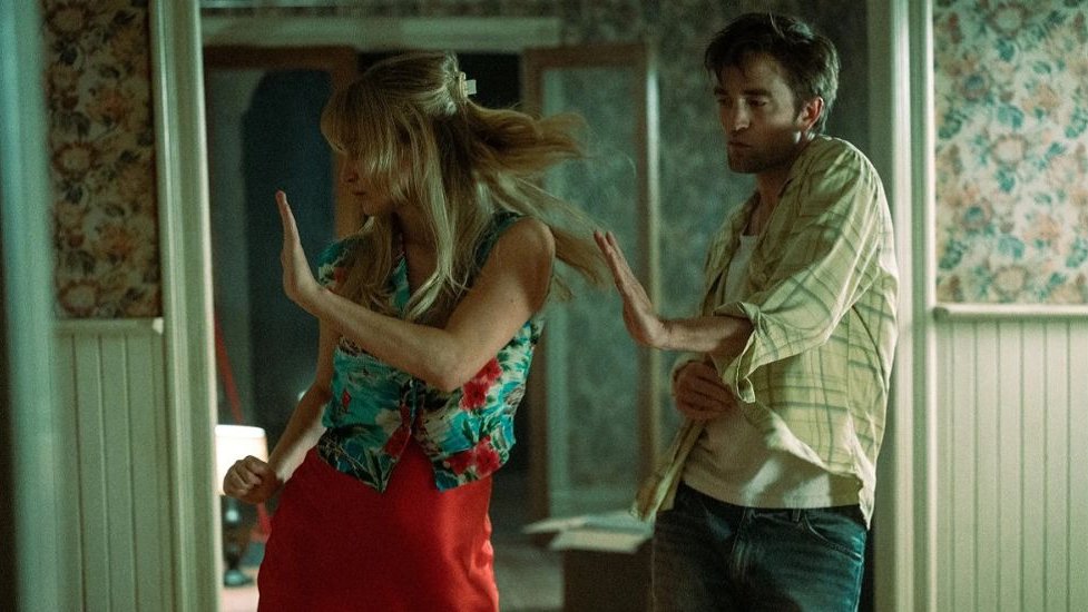 A woman and a man dance side by side. A wallpapered wall in the background.