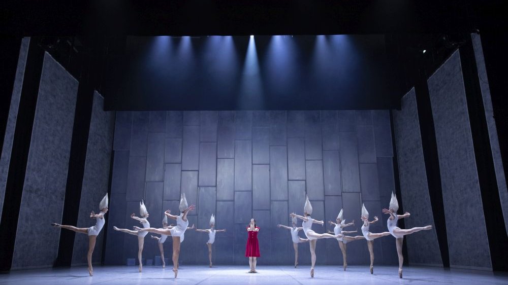 On the stage there are ballerinas in white costumes, and between them in the middle stands a woman in a red dress.