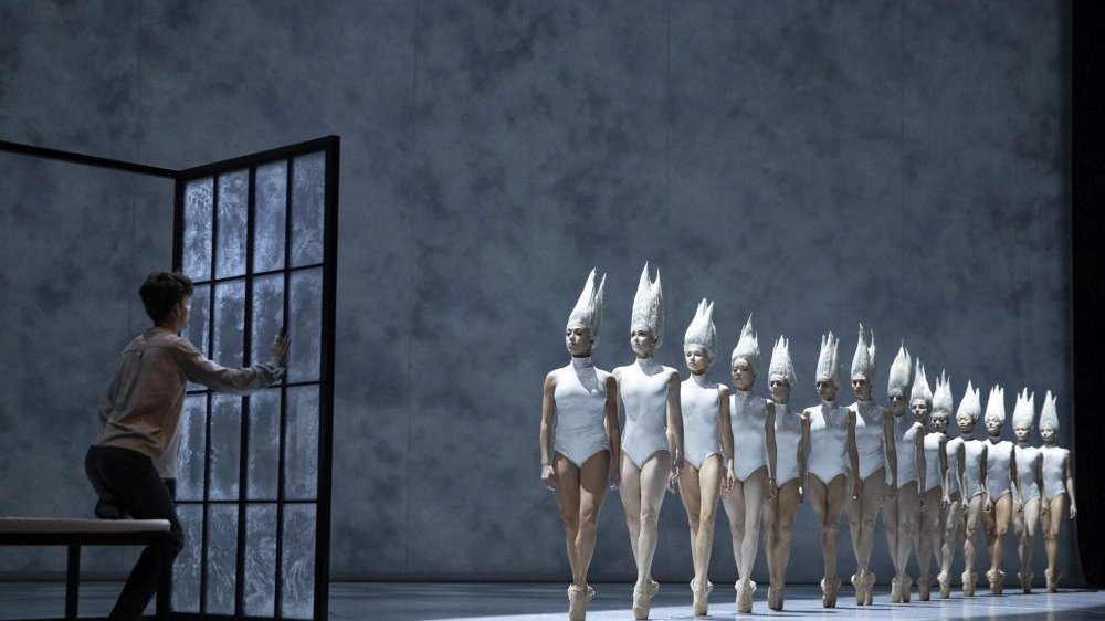 On the stage, a boy stands behind a glass door, and opposite him, ballerinas dressed in white walk in a row.