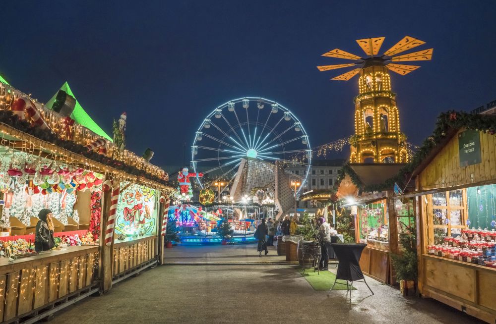 Brightly and colorfully lit stalls with a variety of goods, in the background there is a children's carousel and a Ferris wheel. - grafika artykułu