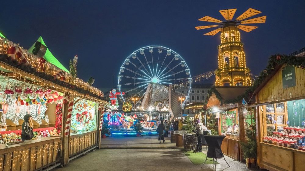 Brightly and colorfully lit stalls with a variety of goods, in the background there is a children's carousel and a Ferris wheel.