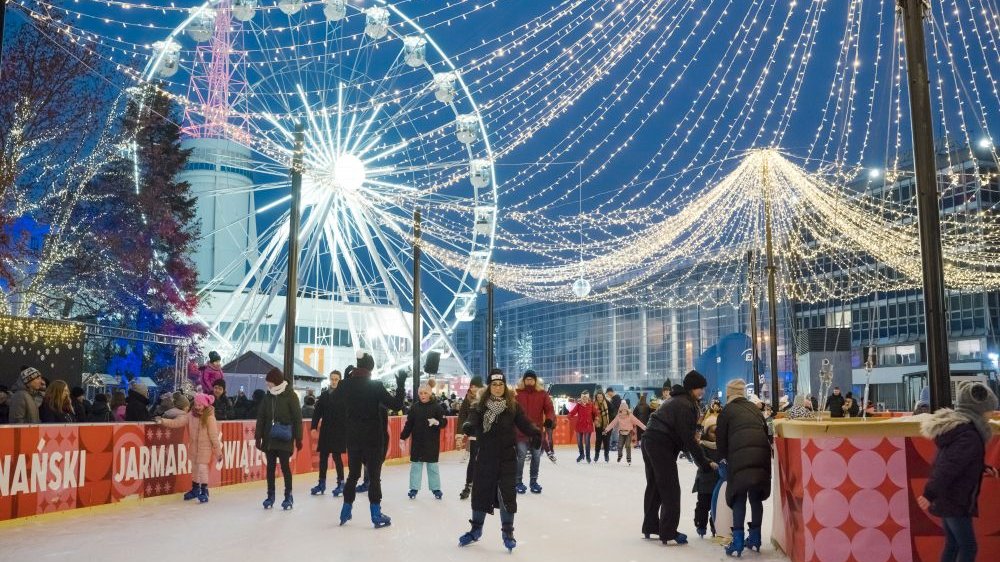 A dozen or so people skate on a small ice rink, above which glowing lights are strung. In the background is the building known as the Iglica at the Poznań International Fair.